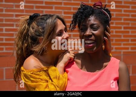Fröhliche multirassische weibliche Freundinnen in farbenfrohen Kleidern, die kuscheln und Spaß haben, während sie den Sommertag gemeinsam gegen die Mauer genießen Stockfoto