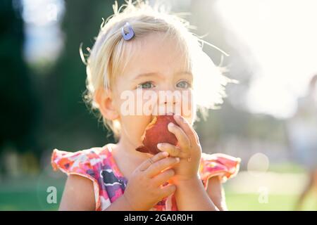 Das kleine Mädchen isst einen Pfirsich auf einem grünen Rasen und hält ihn mit den Händen. Hochformat Stockfoto