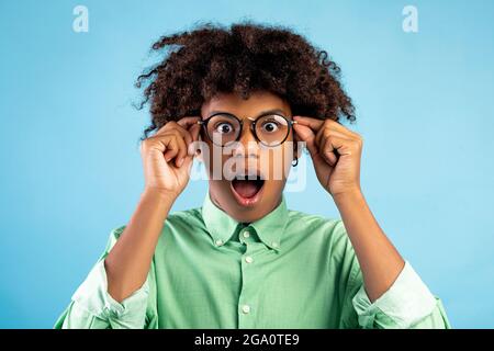 Unglaublich. Schockierter schwarzer Teenager, der seine Brille berührte, seinen Mund öffnete und auf blauem Hintergrund auf die Kamera schaute Stockfoto