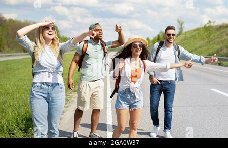 Glückliche multiethnische Freunde versuchen, freie Fahrt zu bekommen, Anhalter auf der Autobahn, zeigen Daumen nach oben Gesten im Freien, Panorama Stockfoto