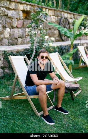 Mann in Sonnenbrille sitzt auf einem Klappstuhl auf einem grünen Rasen in der Nähe einer Steinmauer Stockfoto