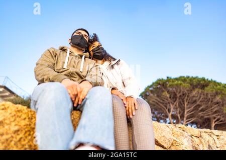 Ein junges, verliebtes multirassisches Paar, das an einer Wand sitzt und eine schwarze, sichere Gesichtsmaske trägt, schaut Kopf an Kopf auf den Horizont bei Sonnenuntergang. Pandemie weltweit Stockfoto