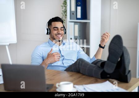 Fröhlicher arabischer Mitarbeiter, der in einem modernen Büro Musik in Kopfhörern hört und virtuelle Gitarre spielt Stockfoto