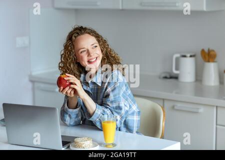 Genießen Sie die Zeit zu Hause. Schöne junge lächelnde Frau, die am Laptop arbeitet und Orangensaft trinkt, während sie zu Hause auf dem Stuhl in der Küche sitzt. Ja Stockfoto