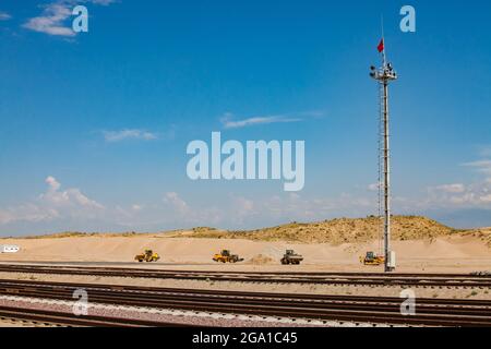 Abstrakter Blick auf Wüste und Sandhaufen. Leuchtenmast, rote Fahne oben. Eisenbahn und Traktoren vorne, blauer Himmel mit hellen Wolken im Hintergrund. Stockfoto