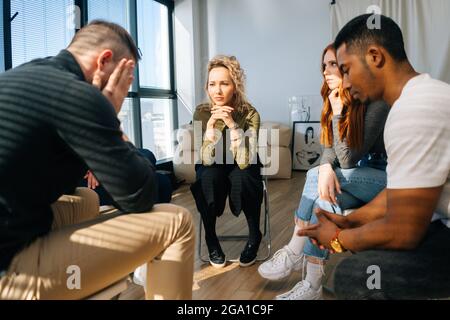 Rückansicht eines traurigen, verzweifelten jungen Mannes, der anderen Patienten im Kreis traurige Geschichten über psychische Probleme oder sucht erzählt. Stockfoto