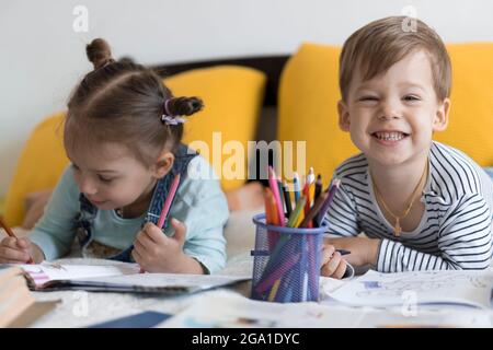 Smart lächelnd Vorschool Kleinkind Kinder Junge und Mädchen zeichnen mit Bleistiften liegen auf dem Bauch am gelben Bett. Kleine niedliche Leser haben Spaß, glücklich Kind auf Stockfoto