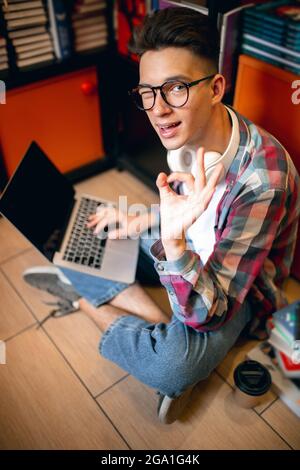 Ein junger Kaukasier, Student, der auf dem Boden mit einem Laptop über dem Haus oder der Bibliothek sitzt. Stockfoto