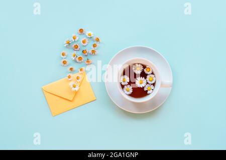 Eine Tasse Kamillentee, ein Umschlag und Gänseblümchen auf blauem Hintergrund. Draufsicht, flach liegend, Kopierbereich. Stockfoto