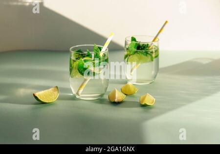 Zwei Gläser Limonade mit Stroh und Limettenscheiben auf blauem Tisch. Kalte erfrischende Getränke gegen weiße Wand im Sonnenlicht. Stockfoto