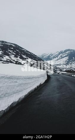 Vertikale Aufnahme einer nassen Asphaltstraße, die von kleinen schneebedeckten Hügeln umgeben ist Stockfoto