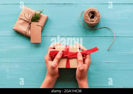 Weihnachtshintergrund mit Geschenkschachteln, Skein aus Seil auf blauem blauem Holztisch. Vorbereitung auf den Urlaub. Draufsicht, flach liegend. Die Hände des Mädchens halten das Geschenk Stockfoto