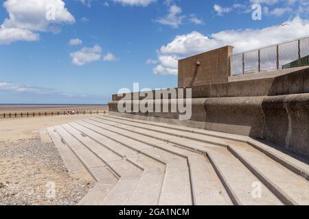 Rhyl North Wales, Blick auf Meer, Sand und Stein, Betonstufen, die als Sitzfläche genutzt werden, um die Welt an sich vorbei zu beobachten. Blauer Himmel mit weißen, flauschigen Wolken Stockfoto