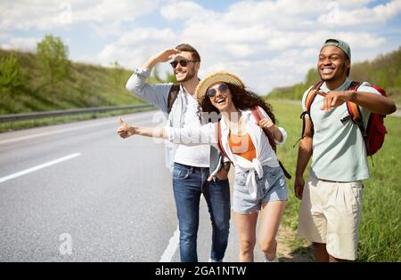 Diverse Freunde stehen am Straßenrand, halten Autos an, trampen für freie Fahrt, genießen Sommerferien Stockfoto