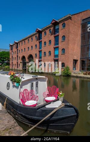 Traditionelles Hausboot-Schmalboot auf dem Kanal im Stadtzentrum von manchester dockt mit zwei Sitzen am Vordereck und alten roten Ziegelmühlen dahinter an. Stockfoto