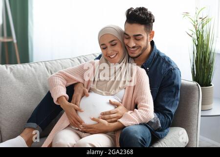 Frohe Erwartungen. Liebevoller arabischer Ehemann umarmt und berührt den Bauch der Schwangeren Stockfoto