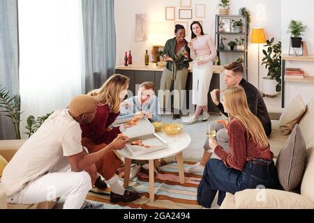 Gruppe von jungen fröhlichen interkulturellen Freunden in Smart Casualwear sitzen am kleinen weißen runden Tisch im Wohnzimmer, mit Pizza und Getränken Stockfoto