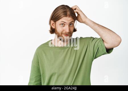 Bild eines hübschen Hipster-Mannes, Mann mit blonden Haaren und Bart, der seinen Kopf schürft und unwissend lächelt, der in einem grünen T-Shirt gegen Weiß steht Stockfoto