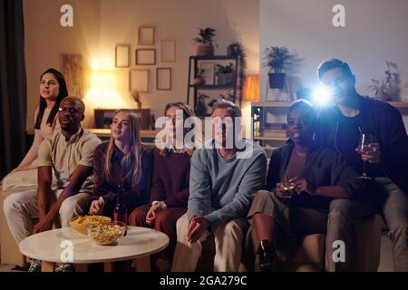 Gruppe junger interkultureller Freunde, die auf der langen Couch sitzen Kleiner Tisch mit Snacks und Film zusammen im Wohnzimmer Auf der Party Stockfoto