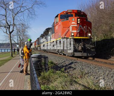 Eine junge Familie steht auf einem Wanderweg an der Küste von British Columbia und beobachtet einen Güterzug, der auf den Gleisen vorbeifährt; White Rock, British Columbia, Kanada Stockfoto