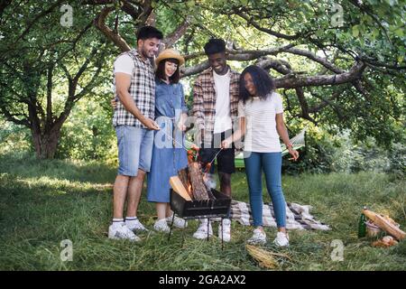 Eine Gruppe von vier multikulturellen Freunden, die sich beim Grillen von Würstchen treffen. Junge Menschen im Sommer Kleidung Kochen Lebensmittel auf Feuer im Freien. Stockfoto