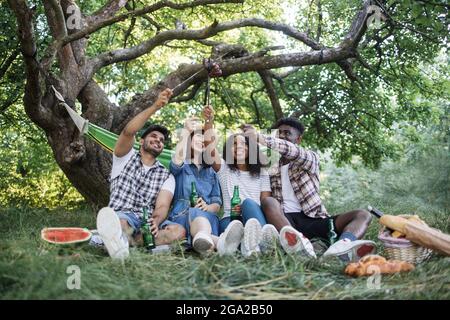 Vier junge Menschen in legerer Kleidung sitzen zusammen im grünen Garten und genießen ein Sommerpicknick. Fröhliche multikulturelle Freunde, die kaltes Bier trinken und gegrillte Würstchen essen. Stockfoto