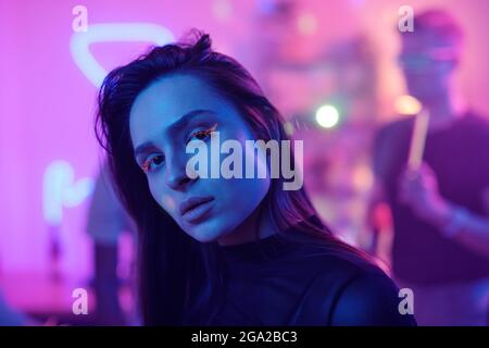 Junge Frau mit glamourösen Neon Make-up Blick auf Sie, während Vor der Kamera stehend gegen ihre tanzenden Freunde und Party genießen Stockfoto