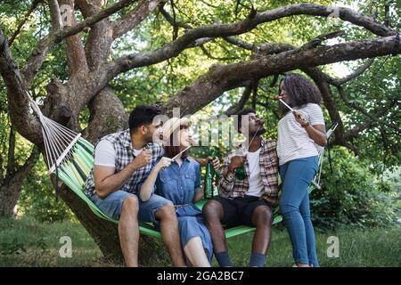 Fröhliche, vielfältige Freunde, die gegrillte Würstchen vom Spieß essen und Bier trinken, während sie im Freien in der Hängematte sitzen. Konzept der Menschen, Sommer Picknick und Entspannung. Stockfoto
