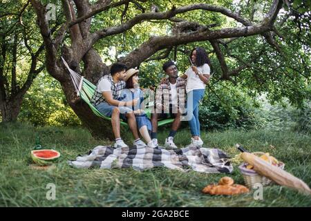 Gruppe von vier multikulturellen Freunden, die in ihrer Freizeit in der Natur essen und Bier trinken. Junge Leute, die im Sommergarten picknicken. Stockfoto