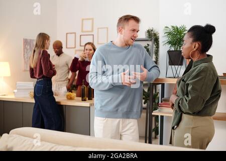 Happy Young liebevolles Paar mit Flaschen Bier Blick auf Sie mit toothy Lächeln während genießen Home Party gegen Chat Freunde Stockfoto