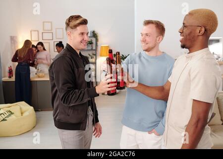 Glückliche junge interkulturelle Männer klirren mit Flaschen Bier und Einander mit toothy Lächeln beim Jubeln angucken Zu Hause Party Stockfoto