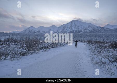 Blick von hinten auf eine Frau, die mit ausgestreckten Armen inmitten einer schneebedeckten Straße steht, in Ehrfurcht vor dem Blick auf die Berge in der ... Stockfoto