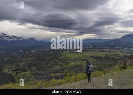 Blick von hinten auf eine Wanderfrau, die auf einem Berghang steht und den Blick auf das Tal und die Bergketten in der Ferne unter ... Stockfoto