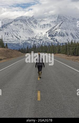 Blick von hinten auf eine Reisende Frau, die mitten auf dem Alaska Highway in Richtung Berge geht, im traditionellen Territorium... Stockfoto