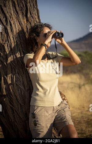 Porträt einer Frau, die sich in kurzen Hosen auf einem Baumstamm lehnt und durch ein Fernglas in das helle Sonnenlicht in die Ferne am Gabus G... Stockfoto