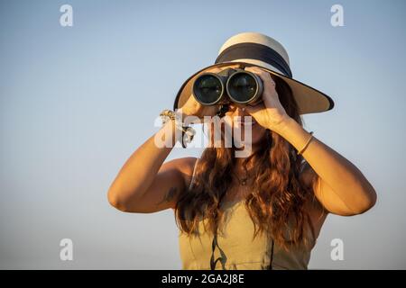 Nahaufnahme einer Frau mit Strohhut und Blick durch ein Fernglas in das helle Sonnenlicht vor einem blauen Himmel auf der Gabus Game Ranch Stockfoto