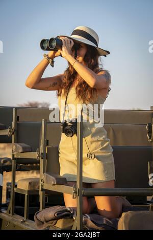 Nahaufnahme einer Frau, die auf einer Safari in einem Jeep kniet, einen Strohhut mit einer Kamera trägt, die um ihren Hals hängt und mit Ferngläsern auf die... Stockfoto