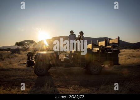 Safariführer im Gespräch mit Gästen, während er in einem Jeep auf der Savanne auf der Gabus Game Ranch bei Sonnenaufgang steht; Otavi, Otjozondjupa, Namibia Stockfoto