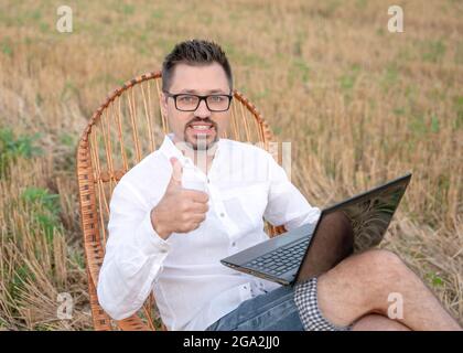 Nahaufnahme eines bärtigen Mannes mit Laptop, der im Freien auf einem Schaukelstuhl sitzt. Glücklicher kaukasischer Mann, der den Daumen hochzeigte und lächelte und von der Empfehlung überrascht war Stockfoto