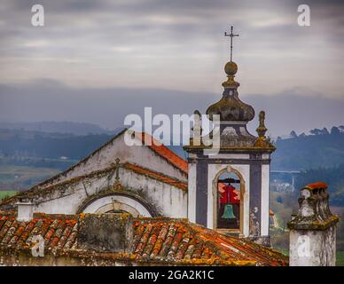 Alter Glockenturm und Ziegeldächer mit Blick auf die Landschaft und die mittelalterliche Stadt Obidos mit einem trüben, bewölkten Himmel Stockfoto