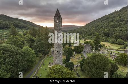 Luftaufnahme des Rundturms, der durch das Fenster auf die Sonne schaut, die über den Hügeln von Glendalough (oder dem Tal der zwei Seen) aufgeht, an der... Stockfoto