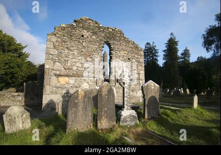 Grabsteine und keltisches Kreuz-Denkmal mit dem runden Turm, der durch das Lanzettenfenster der Ruinen von Glendalough (oder das Tal des Two Lake... Stockfoto