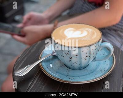 Eine Tasse flaches Weiß oder Cappuccino mit Latte Art serviert in einer Keramikschale mit Untertasse für eine Person, die das Mobiltelefon auf dem Hintergrund hält Stockfoto
