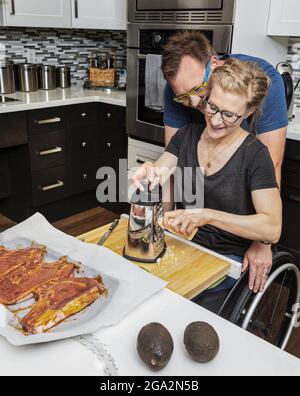 Eine querschnittsgelähmte Frau, die in ihrer Küche ein Essen für ihre Familie zubereitet, während sie von einem Rollstuhl aus arbeitet, als ihr Mann sie umarmt; Edmonton, Alberta, Kanada Stockfoto