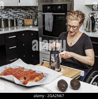 Eine querschnittsgelähmte Frau, die in ihrer Küche ein Essen für ihre Familie zubereitet, während sie von einem Rollstuhl aus arbeitet; Edmonton, Alberta, Kanada Stockfoto