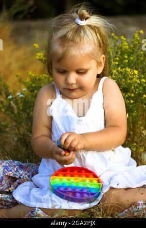 Ein hübsches, kleines blondes Mädchen in einem weißen Kleid spielt im Sommer mit einem Pop auf einer Wiese. Helles Silikon Entwicklung Anti-Stress beliebt Stockfoto