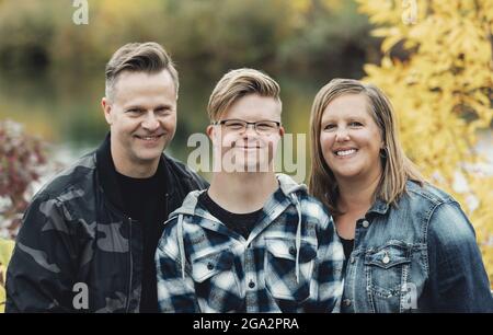 Ein junger Mann mit Down-Syndrom posiert mit Vater und Mutter für ein Familienportrait, während er sich in einem Stadtpark auf einem... Stockfoto