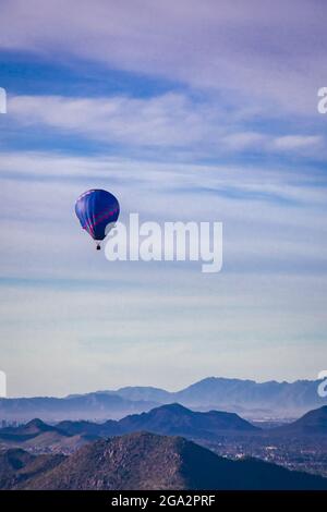 Blau und violett gemusterter Heißluftballon während der Flugabfahrt über die silhouettierten Berge vor einem wolkigen, blauen Himmel Stockfoto