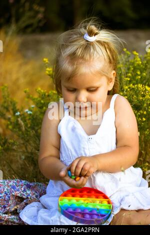 Ein hübsches, kleines blondes Mädchen in einem weißen Kleid spielt im Sommer mit einem Pop auf einer Blumenwiese. Helles Silikon Entwicklungs-Anti-Stress Stockfoto