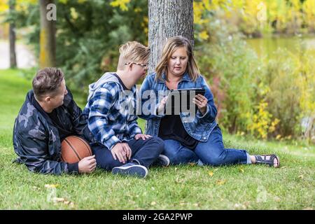 Ein junger Mann mit Down-Syndrom sitzt mit seiner Familie und spielt auf einem Tablet, während er sich in einem Stadtpark auf einem warmen ... Stockfoto